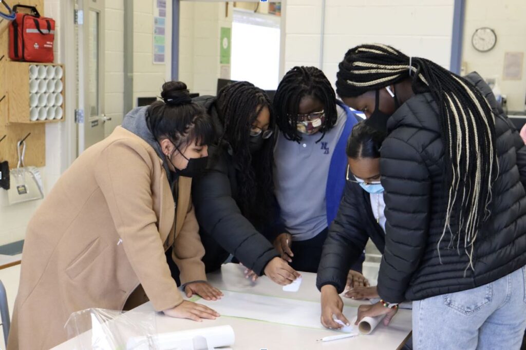 Students standing around a table taking notes on a piece of chart paper.